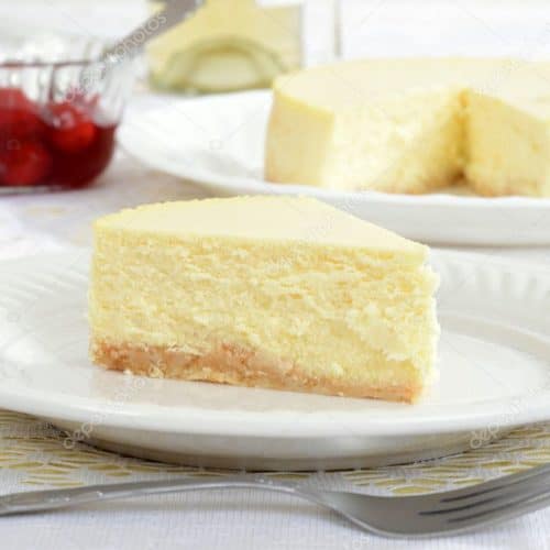 A slice of New York style cheesecake on a white plate with a fork and the rest of the cheesecake in the background.