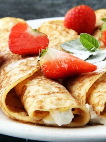 Three strawberry creamy crepes on a white plate with strawberries on top.