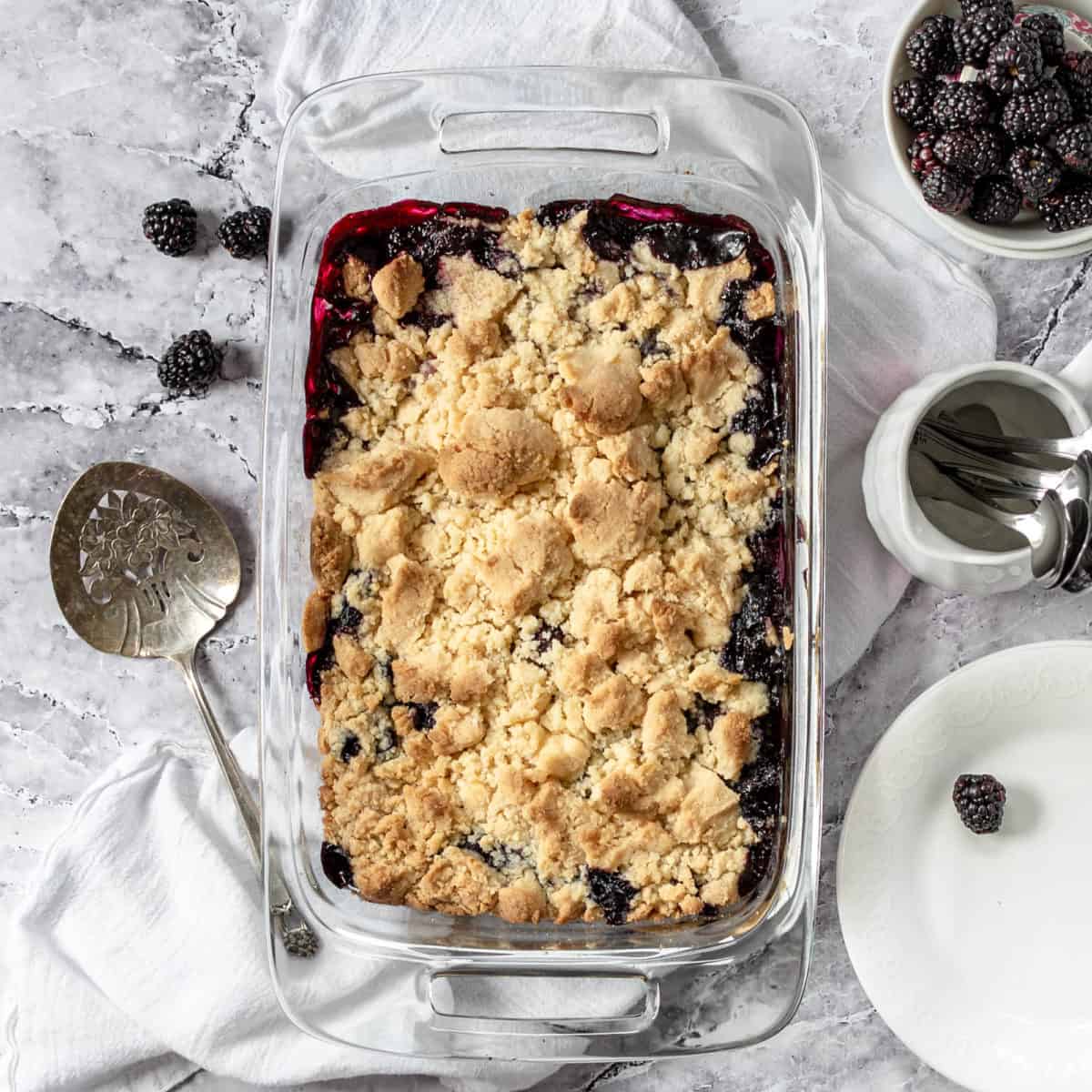 Finished blackberry cobbler dessert in a baking dish with spoons and blackberries.