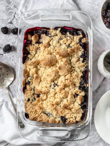 Finished blackberry cobbler dessert in a baking dish with spoons and blackberries.