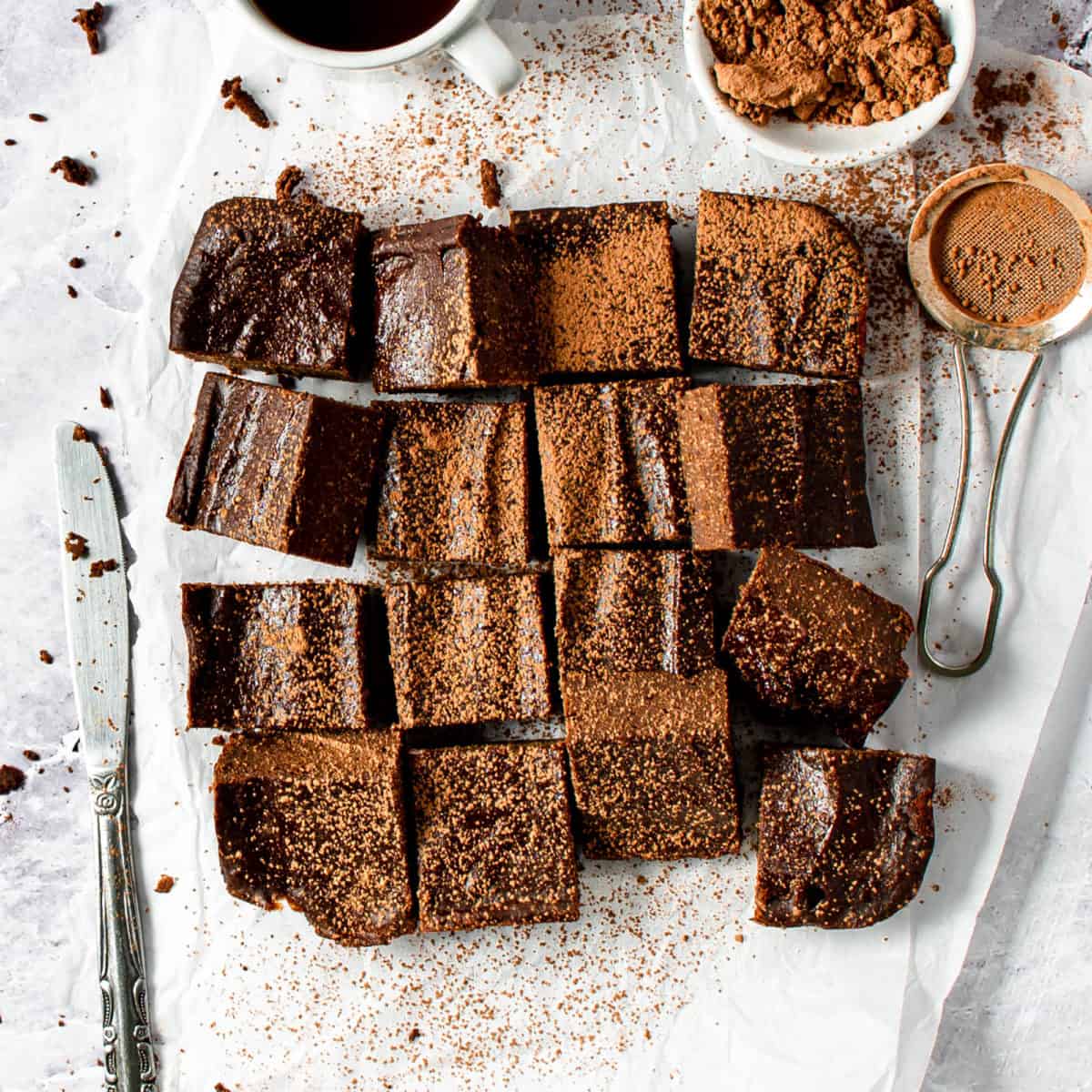 Sixteen brownie squares laid out on a table with a knife and cocoa powder.