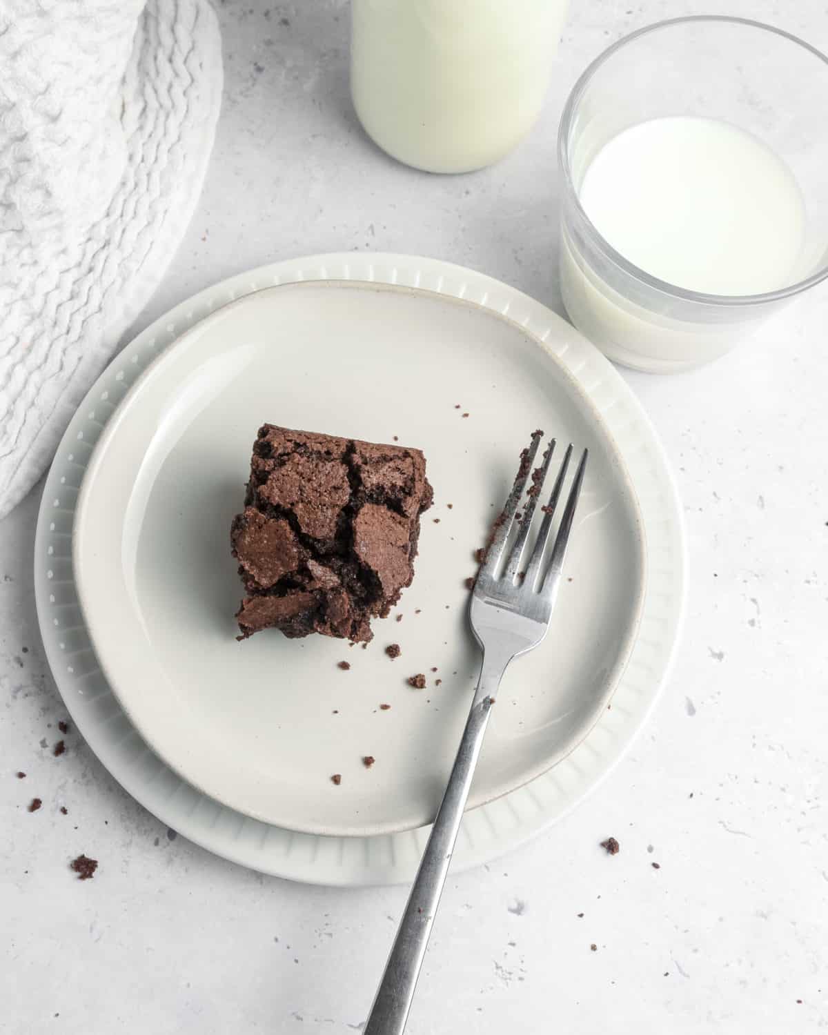 A brownie on a plate surrounded by crumbs and a glass of milk.