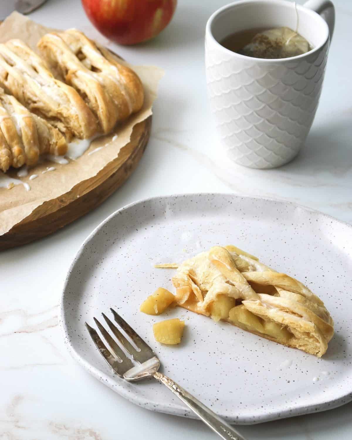 A slice of apple danish on a white plate with a fork and a cup of tea.