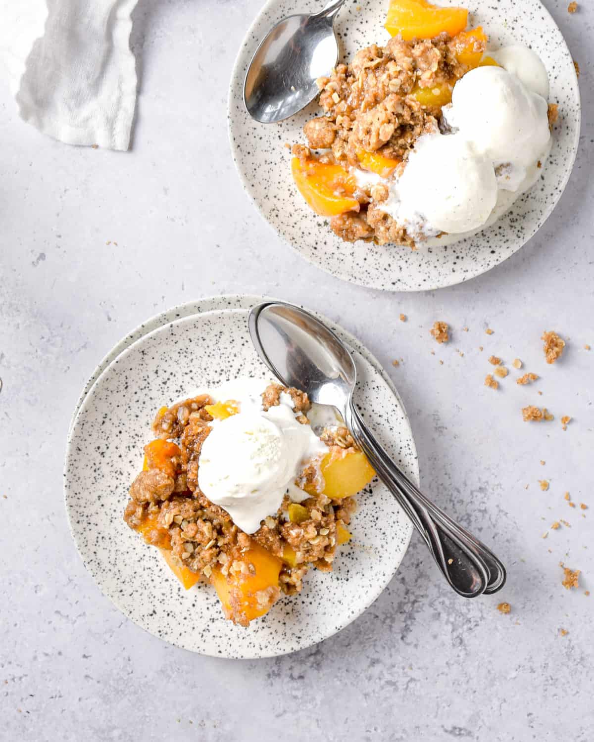 Two plates of peach crisp with spoons and vanilla ice cream.