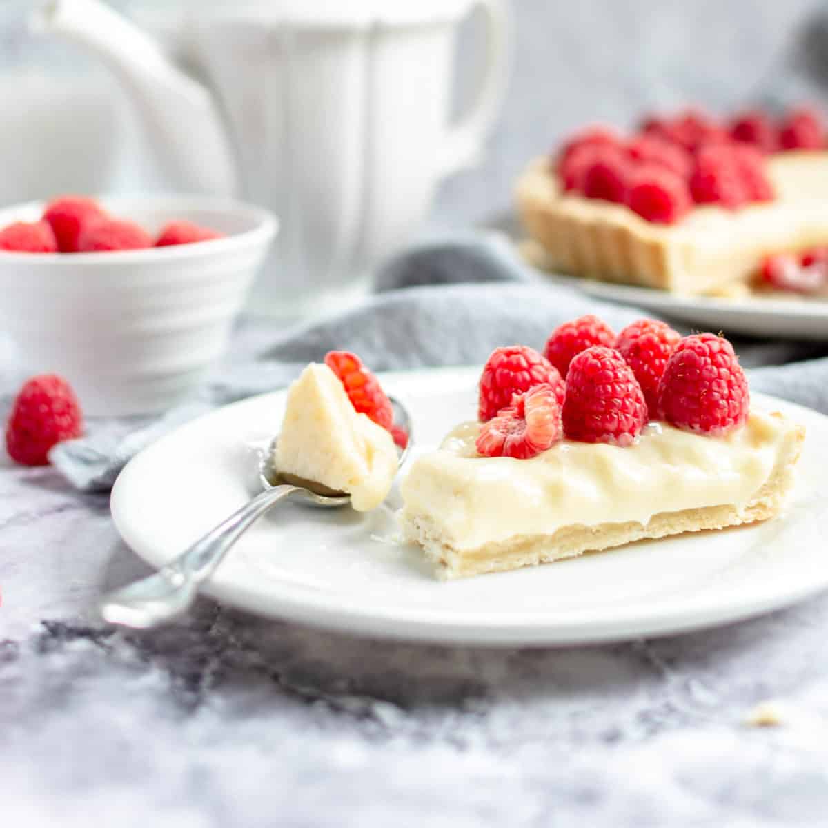 A slice of custard tart on a plate with a spoon and fresh raspberries.
