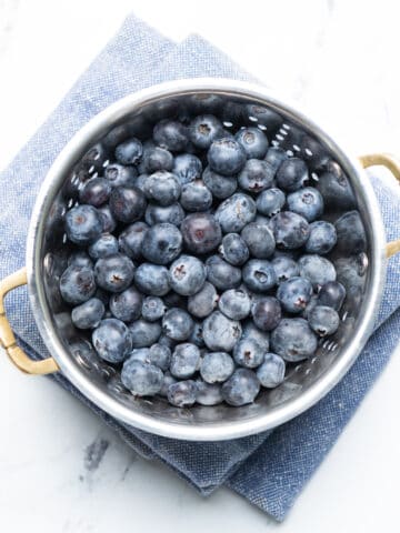 Blueberries in a colander with a blue kitchen towel underneath.
