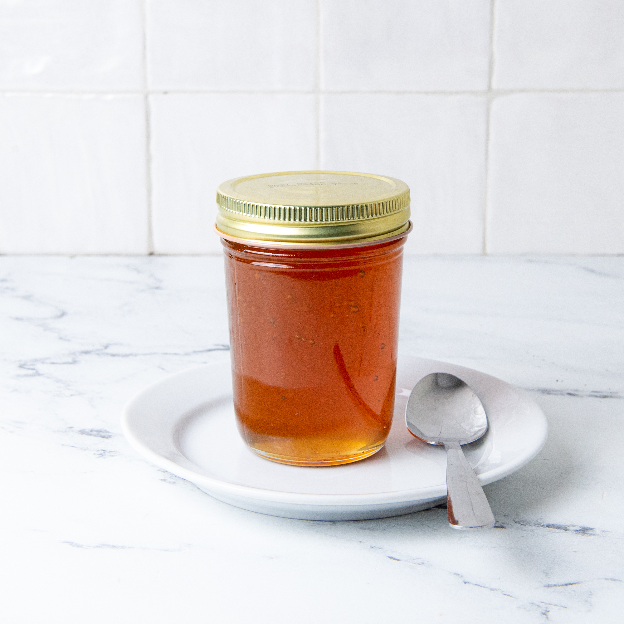 Honey in a clear jar, on a white saucer with a spoon.