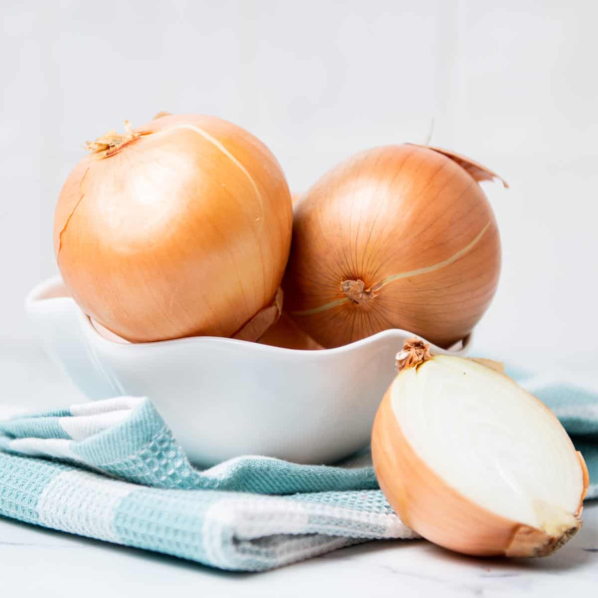Onions in a white bowl on a white and blue kitchen towel with a single sliced onion.