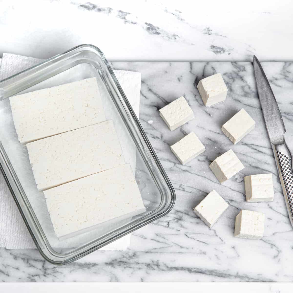 Cutting tofu into cubes from a glass dish and on a kitchen counter.