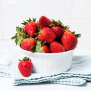 Strawberries in a white bowl, with one on a kitchen cloth.