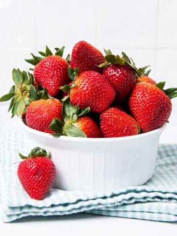 Strawberries in a white bowl, with one on a kitchen cloth.